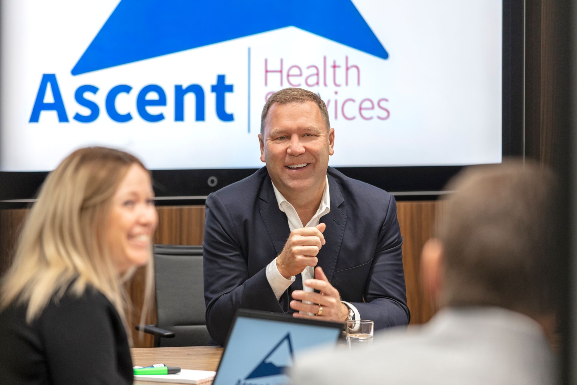 Eine Gruppe von Menschen sitzt an einem Tisch vor einem Bildschirm mit der Aufschrift „Ascent Health Services“