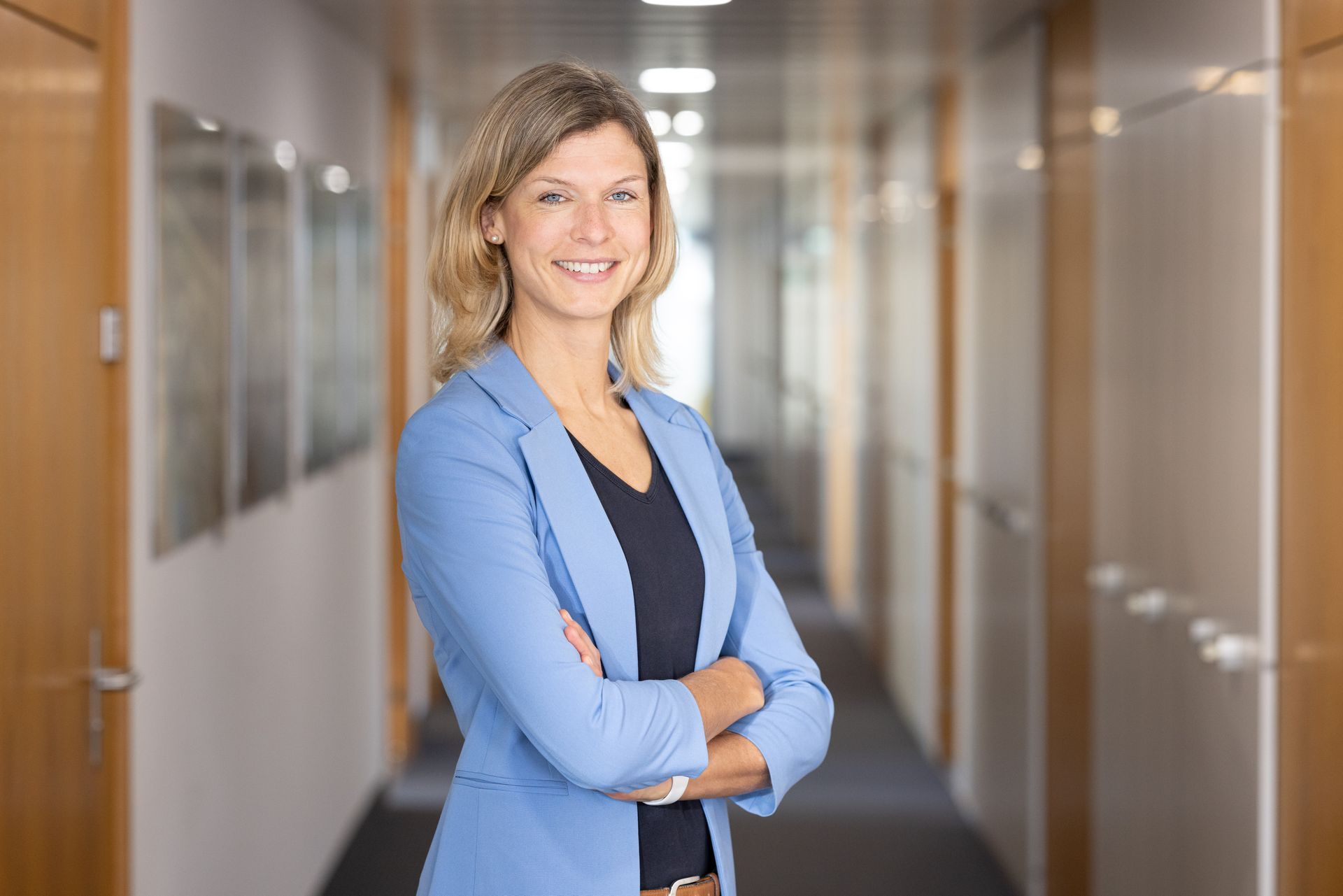 Eine Frau in einer blauen Jacke steht mit verschränkten Armen in einem Flur.