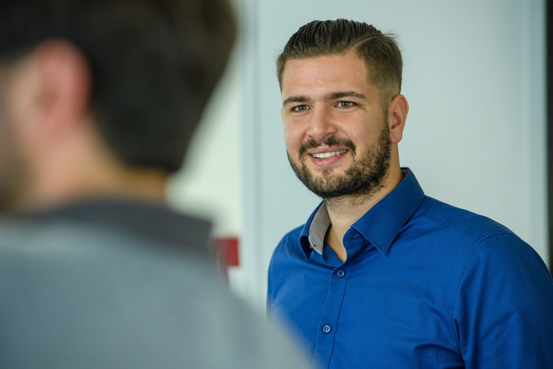 A man in a blue shirt is smiling while talking to another man.