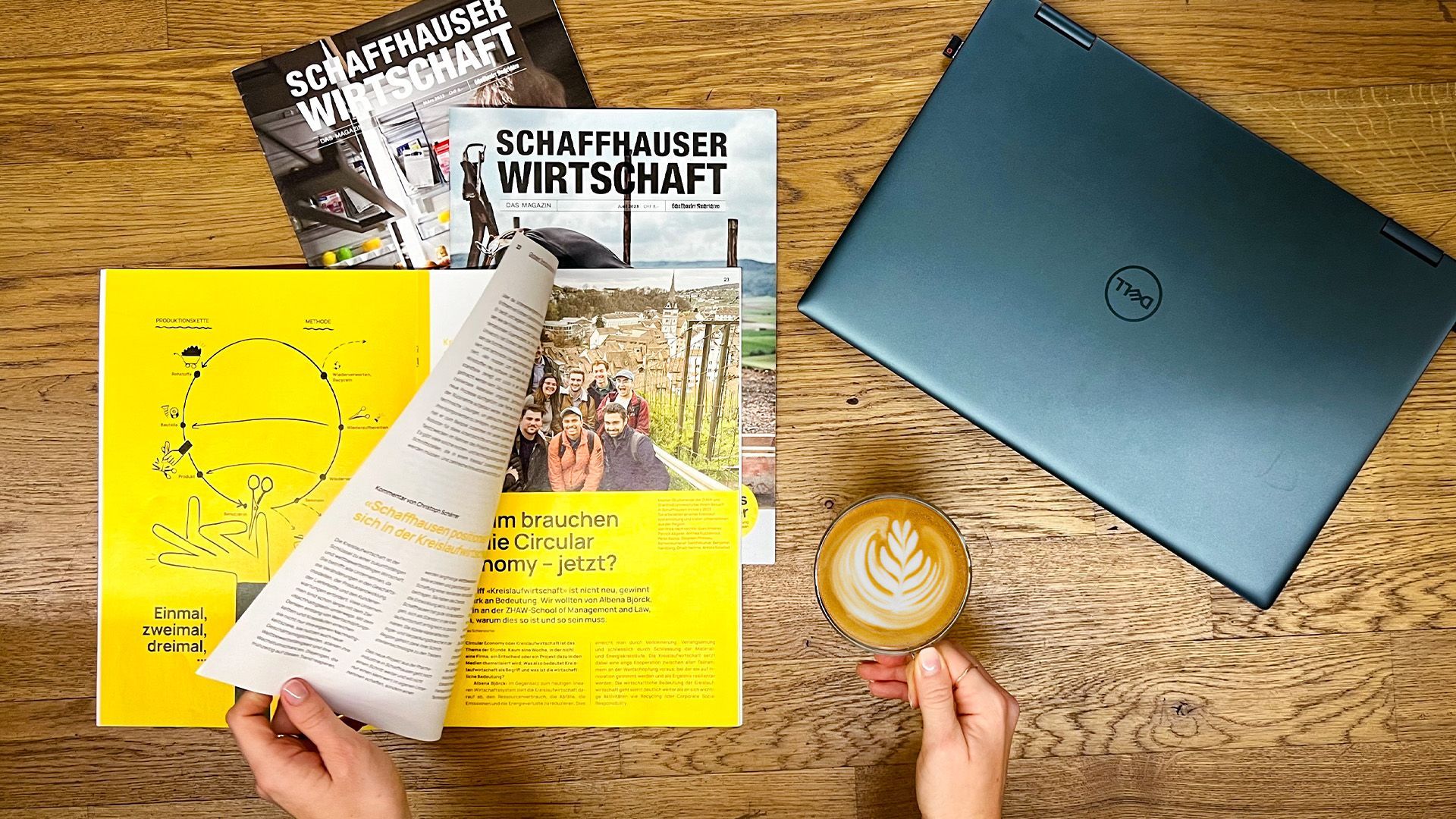 Eine Person hält eine Tasse Kaffee neben einem Laptop und einer Zeitschrift.