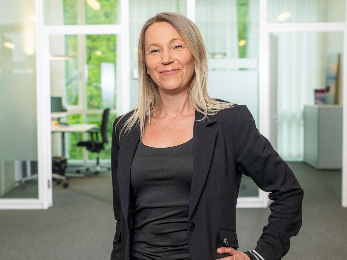 Eine Frau in einer schwarzen Jacke steht mit den Händen in den Hüften in einem Büro.