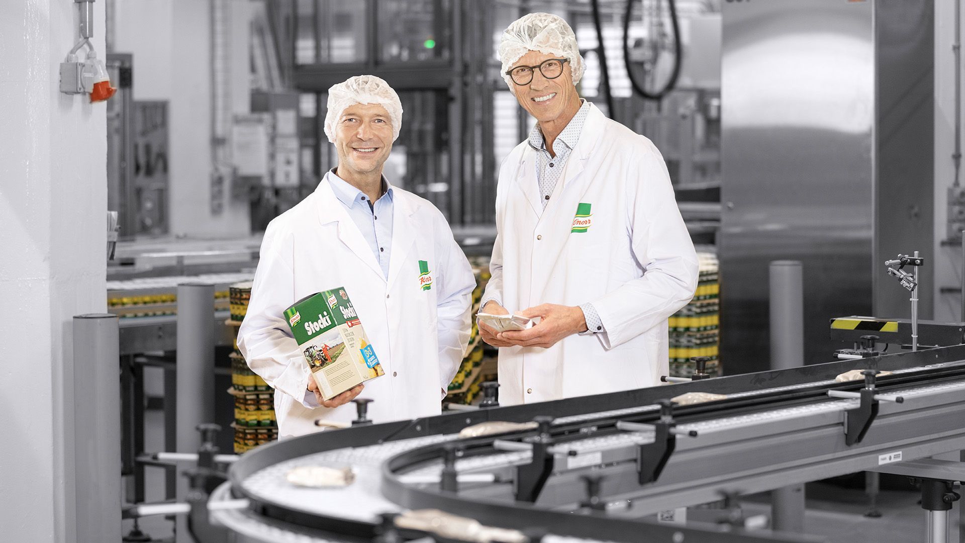 Zwei Männer in Laborkitteln stehen nebeneinander in einer Fabrik.